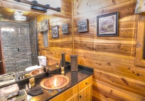 Een houten badkamer met een douche van natuursteen, een koperen wastafel en lijstwerken aan de wanden.