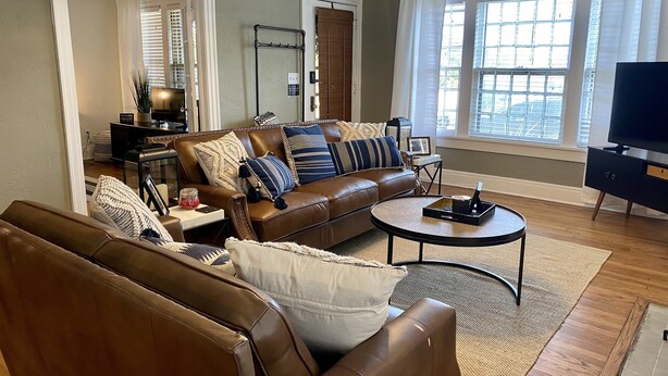 A living room with a brown leather sofa, a coffee table, and a TV. There is a window with blinds and a door with a mirror.