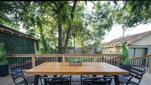 An outdoor wooden deck with a rectangular table and four chairs, surrounded by a patterned rug and a metal railing.