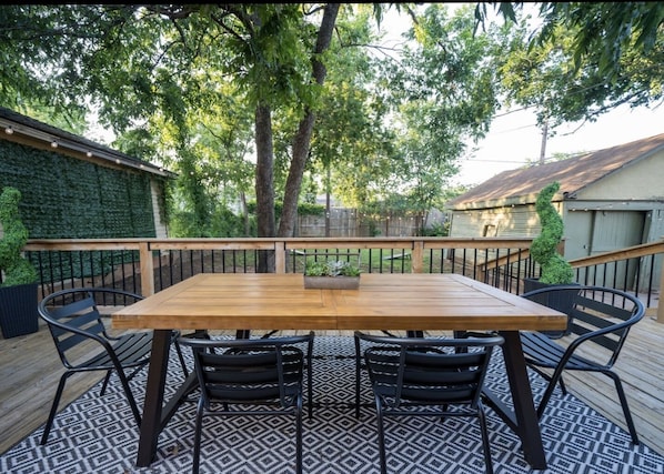 Une terrasse extérieure en bois, avec une table rectangulaire et quatre chaises, entourée d’un tapis à motifs et d’une rambarde en métal.