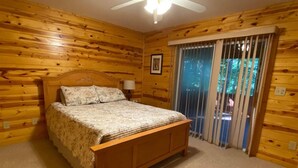 A bedroom with a wooden floor, a bed with a floral bedspread, a ceiling fan, and a window with blinds.