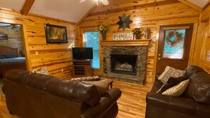 A cozy log cabin interior with a fireplace, two leather sofas, a ceiling fan, and wooden walls.