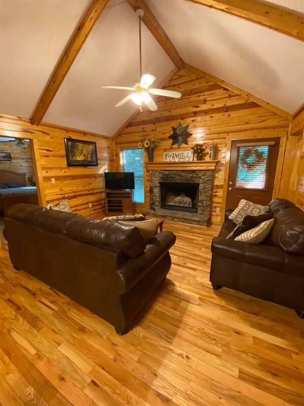 A cozy log cabin interior with a fireplace, two leather sofas, a ceiling fan, and wooden walls.