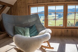 Een knus interieur met hout, een schommelstoel met een bekleed voetenbankje, en een groot raam met uitzicht op een groen landschap.
