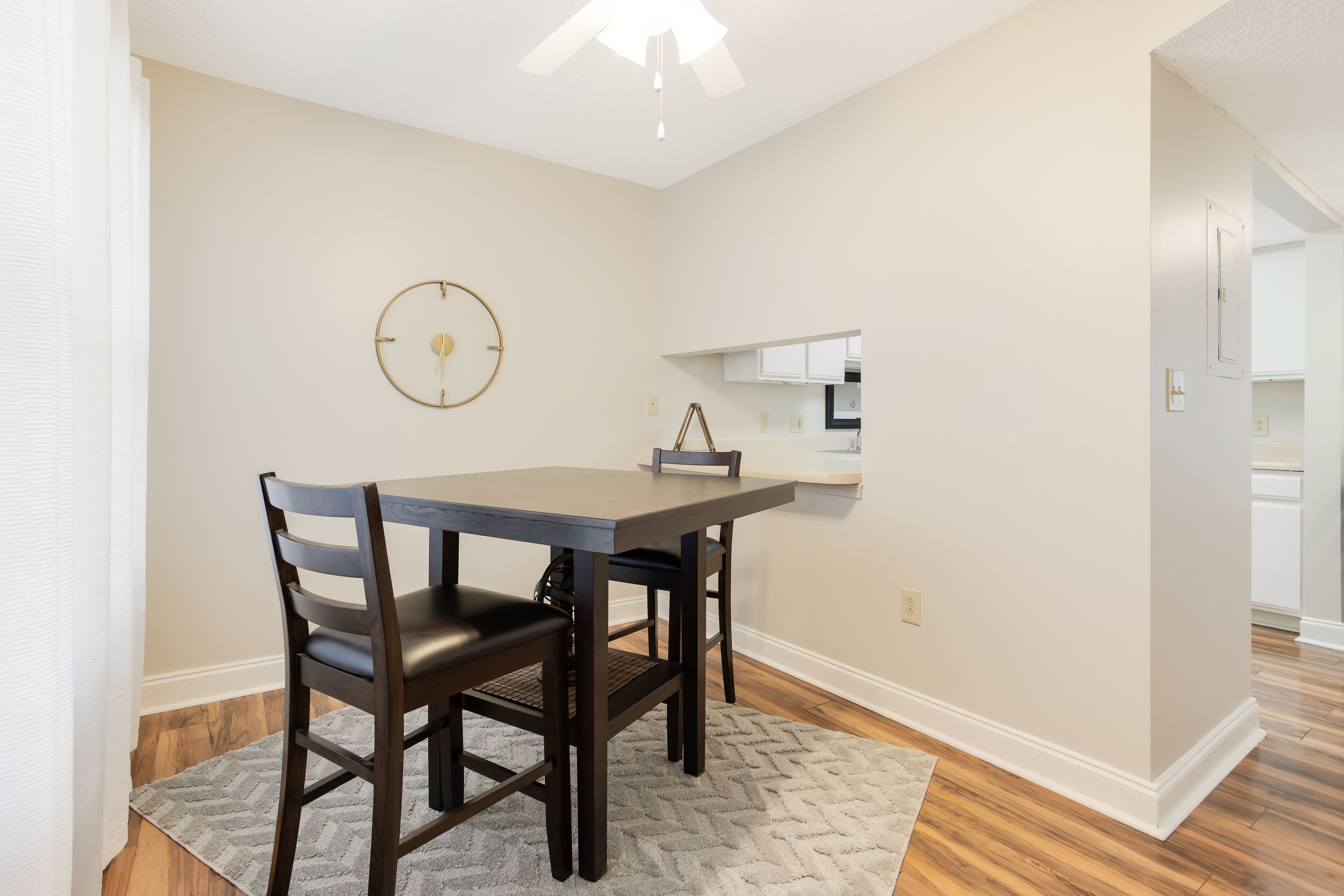A dining area with a table and chairs, a clock on the wall, and a kitchen in the background.