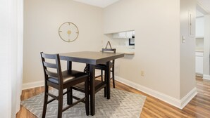 Un coin repas avec une table et des chaises, une horloge au mur et une cuisine en arrière-plan.
