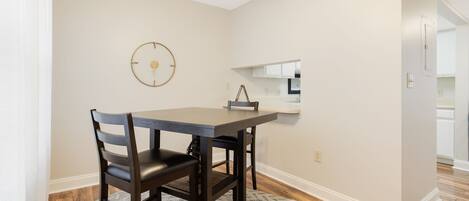 A dining area with a table and chairs, a clock on the wall, and a kitchen in the background.