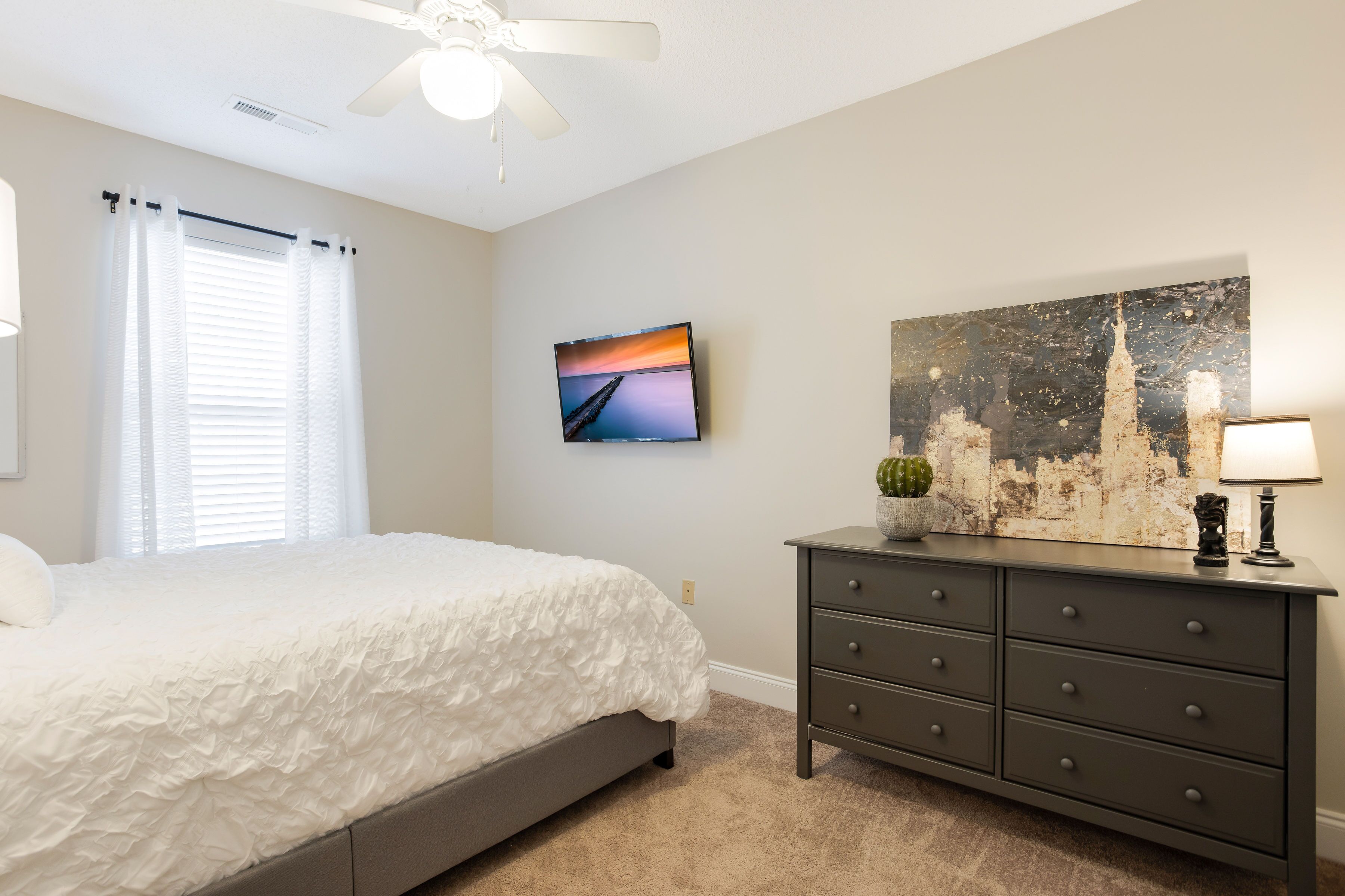 A bedroom with a bed, a dresser, a ceiling fan, a TV, and a painting on the wall.