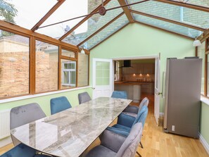 A conservatory with a dining table and chairs, a kitchen visible through an open door, and a large refrigerator. - The Limes (Cambridge)
