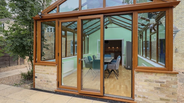 Une véranda dotée d’un toit en verre, d’une structure en bois et offrant une vue sur un espace de restauration intérieur avec une table et des chaises.
