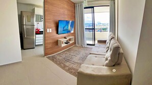 A modern living room with a sofa, a flat-screen TV, a kitchen area with a refrigerator and microwave, and a balcony with a city view. - Vacanze - Smart (Maceió)