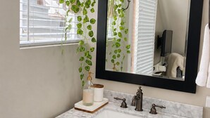 Ein Badezimmer mit einem weißen Marmorwaschbecken, schwarzen Schränken, einem Spiegel und einer hängenden Pflanze.