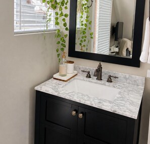Une salle de bain avec un lavabo en marbre blanc, des meubles noirs, un miroir et une plante suspendue.