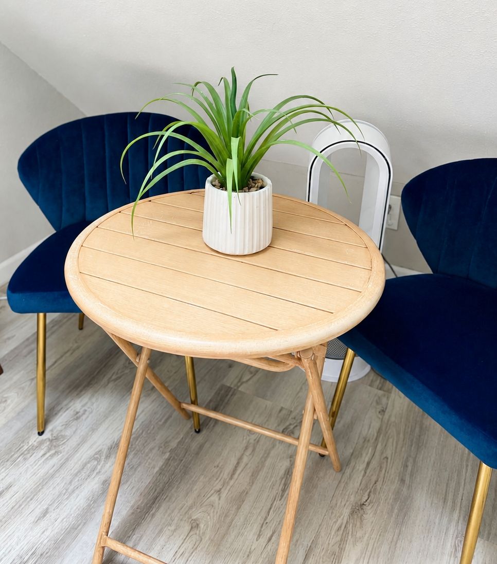 A round wooden table with a white pot containing a plant, flanked by two blue chairs with gold legs, on a light-colored wooden floor.