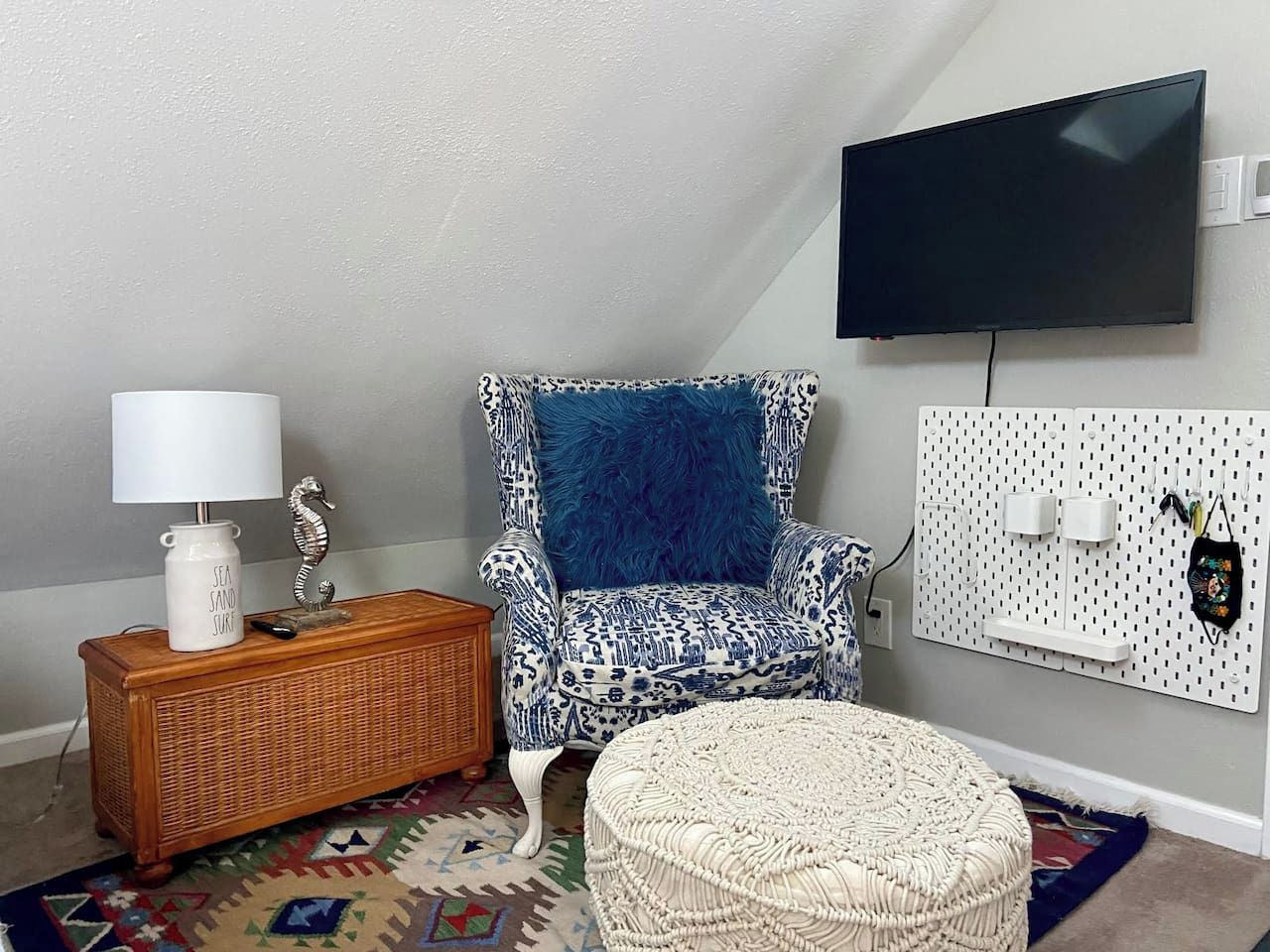 Una habitación con un televisor montado en la pared, un sillón estampado con un cojín azul, una mesita de noche de madera con una lámpara y un otoman tejido.