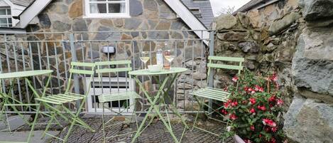 A stone building with a triangular roof, a small window, and a door. There are two green metal chairs and a small round table with a bottle and a glass. A potted plant with red flowers is visible.