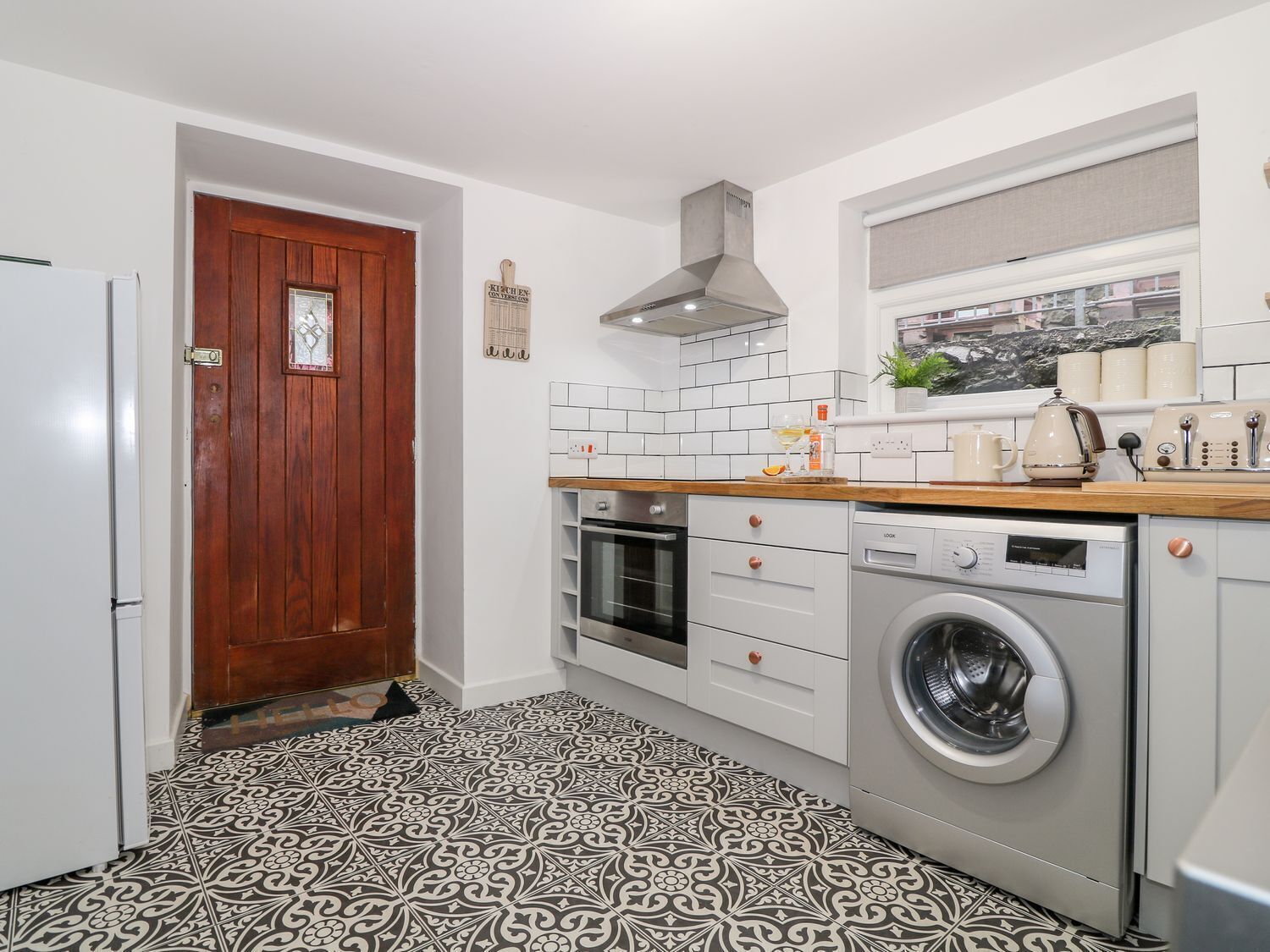 Een kleine keuken met een houten deur, witte betegelde spatwand, een ingebouwde oven, een wasmachine en een raam boven de gootsteen.