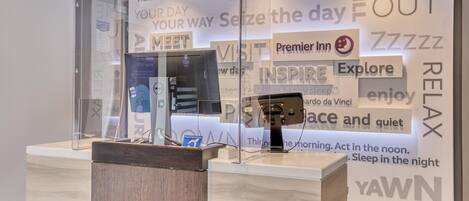 A hotel reception area with a glass partition, a wooden reception desk, and a wall with motivational phrases and the Premier Inn logo.