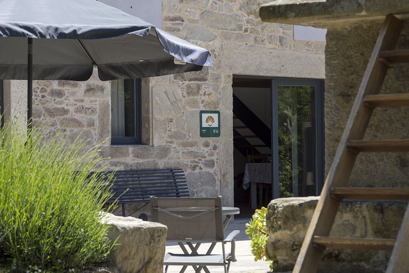 Un bâtiment en pierre avec un banc en bois, une table d’extérieur et un parasol. Un panneau avec un logo est visible sur le mur.