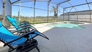 Área externa coberta com cadeiras de descanso azuis, uma piscina e vista para palmeiras e uma cerca.