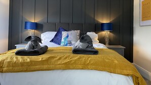 Un lit bien fait, recouvert d’une couvre-lit jaune, avec deux serviettes pliées et un coussin décoratif orné d’un motif de paon. On y trouve deux lampes de chevet avec des abat-jour bleus et un menu encadré accroché au mur.