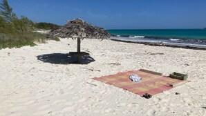 A beach with a thatched umbrella, a blanket, and a green basket on the sand.
