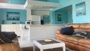 A living room with a brown leather sofa, a white coffee table, and a kitchen area with white cabinets and appliances.