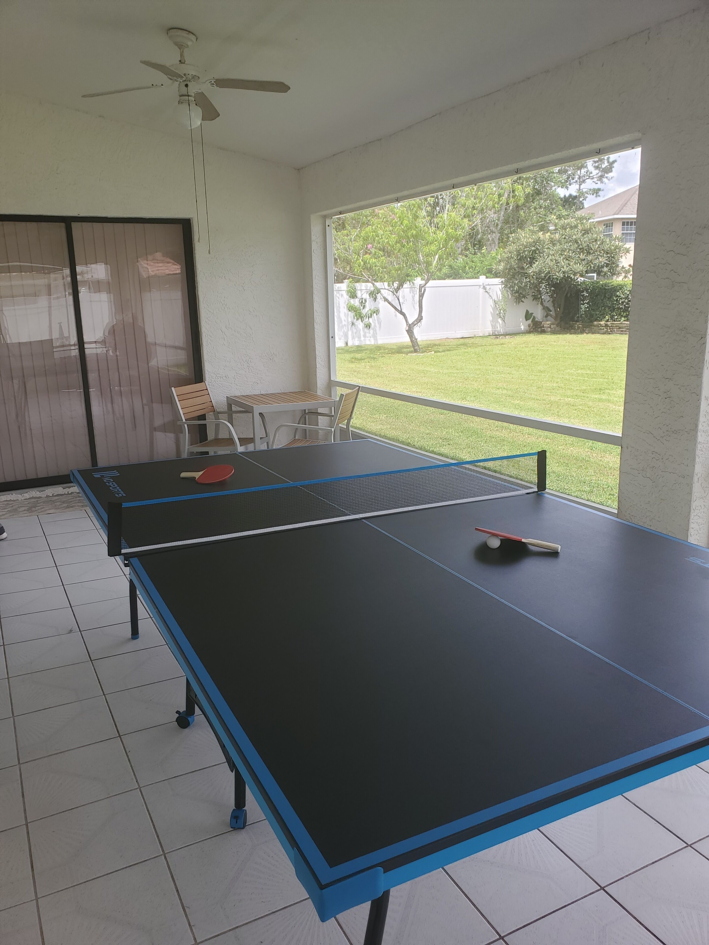 Een tafeltennistafel met stoelen en een tafeltennisracket in een kamer met een plafondventilator, grote ramen en uitzicht op een gazon.