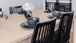 Un coin repas avec une table de couleur claire, des chaises noires et des suspensions blanches. Des tableaux encadrés ornent le mur, et un vase rempli de fleurs est posé sur la table.