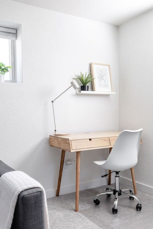 A minimalist office space with a wooden desk, a white office chair, a floor lamp, and a small shelf with a plant and a framed picture.