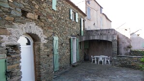 A stone building with a white door and green shutters, a stone archway, and a table with chairs in the courtyard. - Studio (Canari)