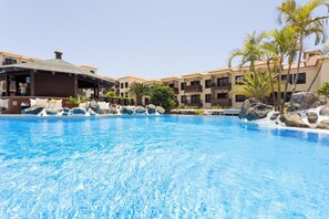 A swimming pool with clear blue water, surrounded by rocks and palm trees, with a building in the background.