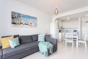 Een moderne woonkamer met een grijze bank, een eettafel met stoelen, een keuken met witte kasten en een hanglamp.