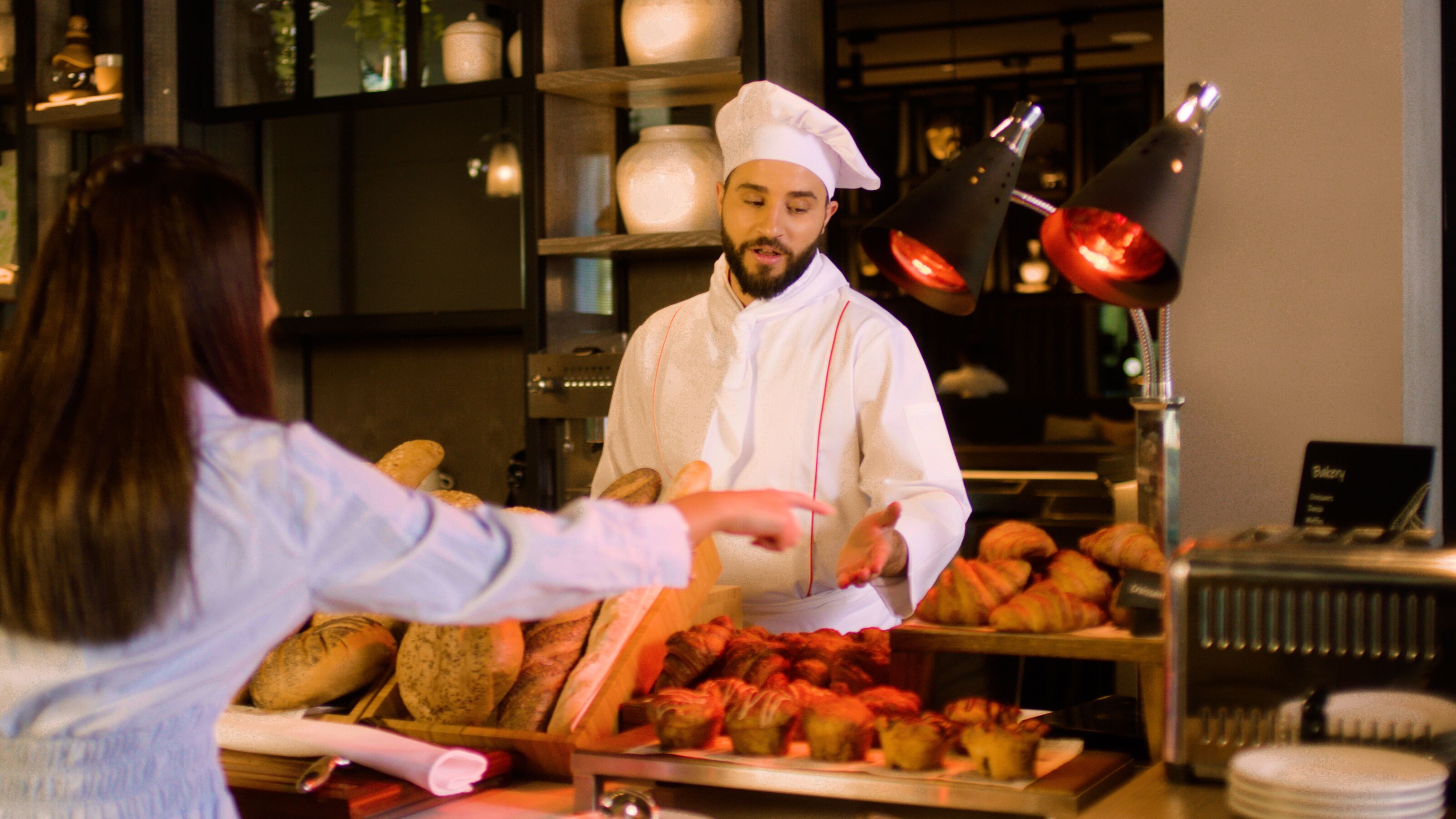 A chef in a white uniform with a chef's hat, serving food to a customer. There are various baked goods on display, including bread and pastries.