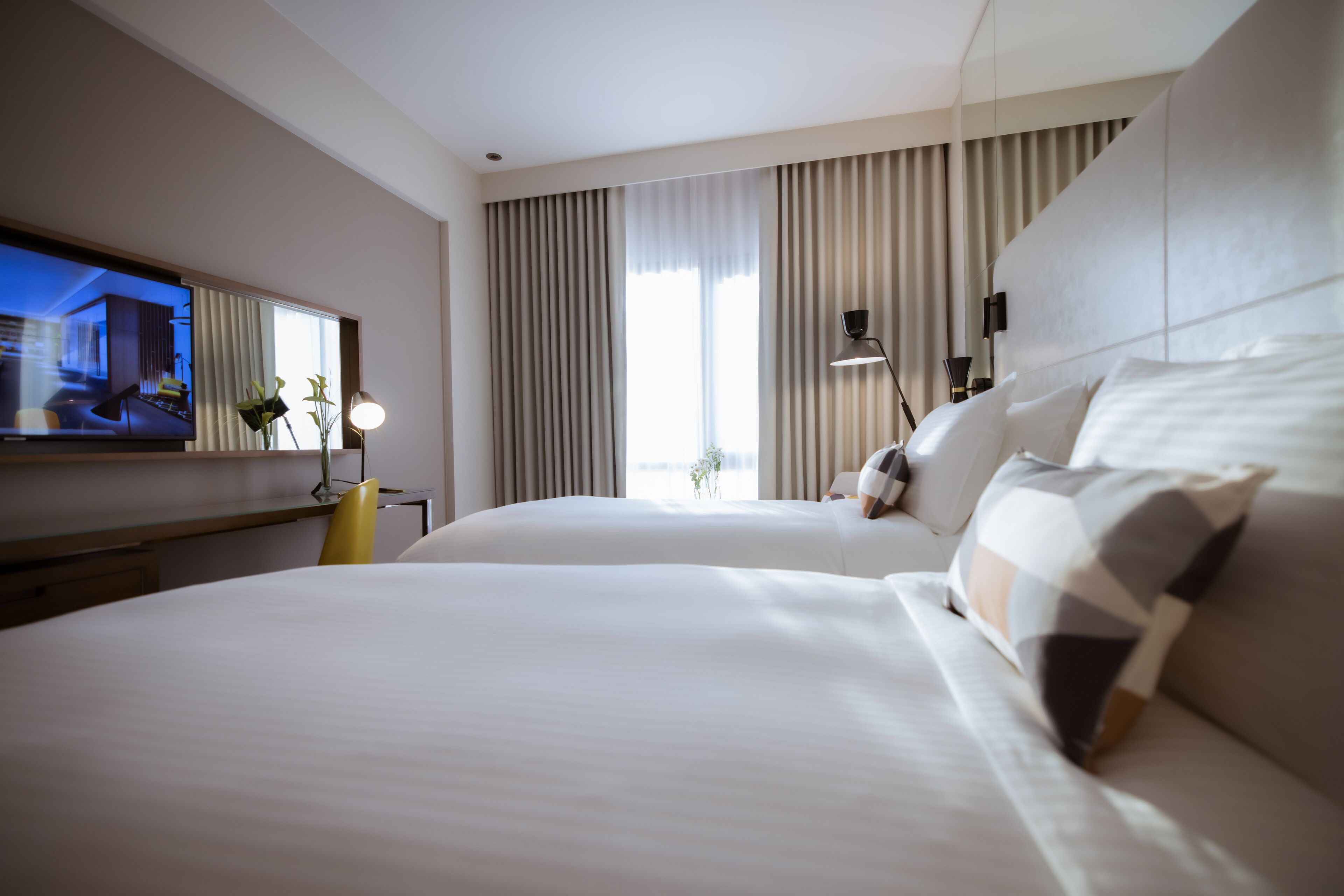 A hotel room with two beds, a TV, a desk, and a large window with curtains.