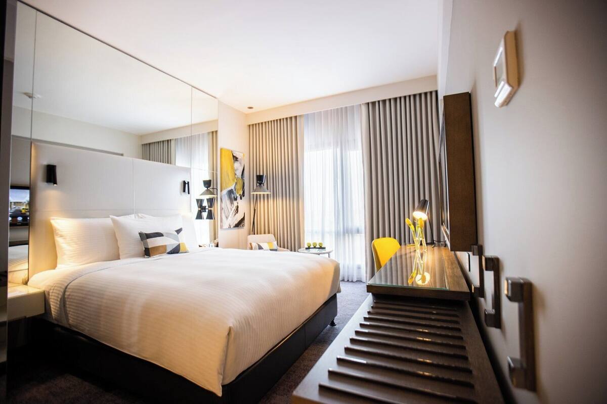 Une chambre d’hôtel moderne avec un grand lit, un bureau et une chaise. On y trouve une fenêtre avec des rideaux, un tableau au mur et une lampe sur le bureau.