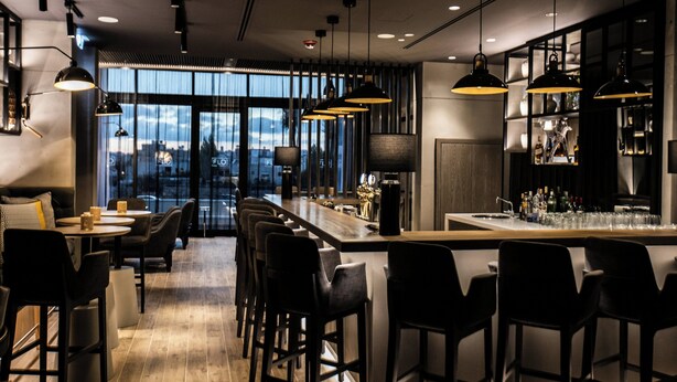 A modern bar area with high stools, pendant lighting, and a city view through large windows. - Centro Mada Amman By Rotana (Amman)
