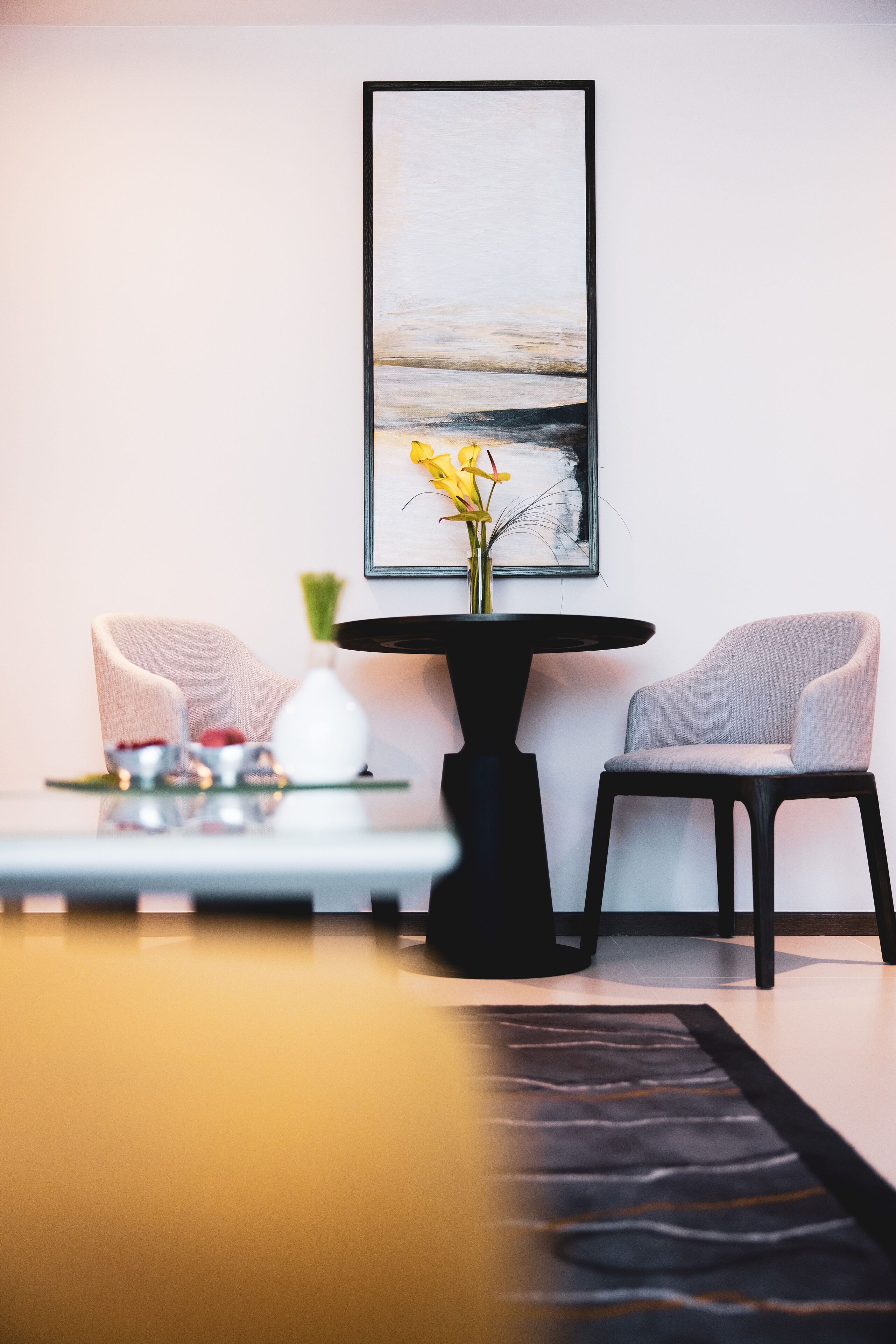 A modern living room with a minimalist design, featuring two grey armchairs, a black coffee table with a vase of yellow flowers, and a framed abstract painting on the wall.