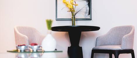 A modern living room with a minimalist design, featuring two grey armchairs, a black coffee table with a vase of yellow flowers, and a framed abstract painting on the wall.