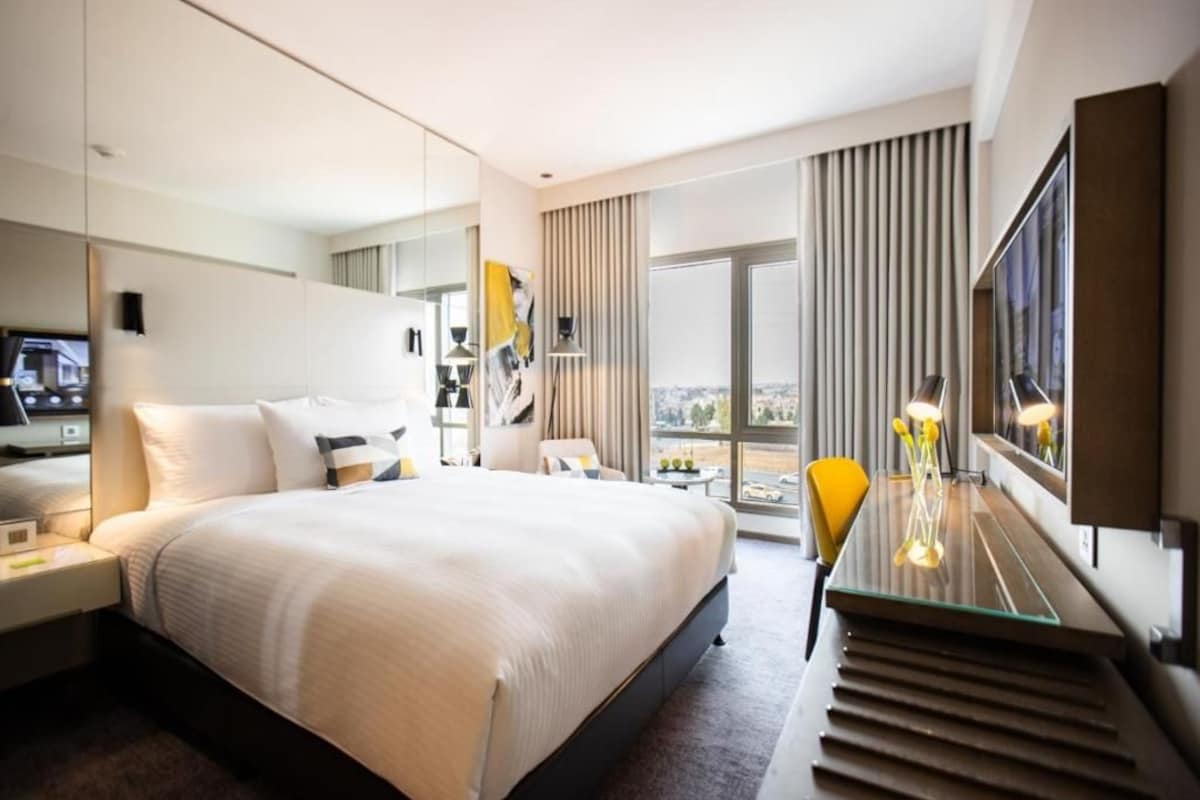 A modern hotel room with a large bed, a desk with a lamp, a chair, a TV, and a large window with a city view.