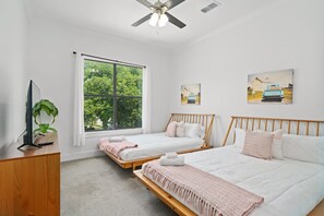 A hotel room with two single beds, a wooden headboard, a ceiling fan, a large window with curtains, a desk with a plant, and artwork on the walls.