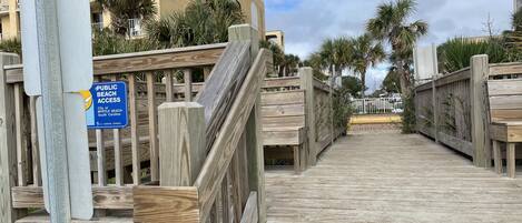 A wooden boardwalk leading to a building with balconies, surrounded by palm trees.