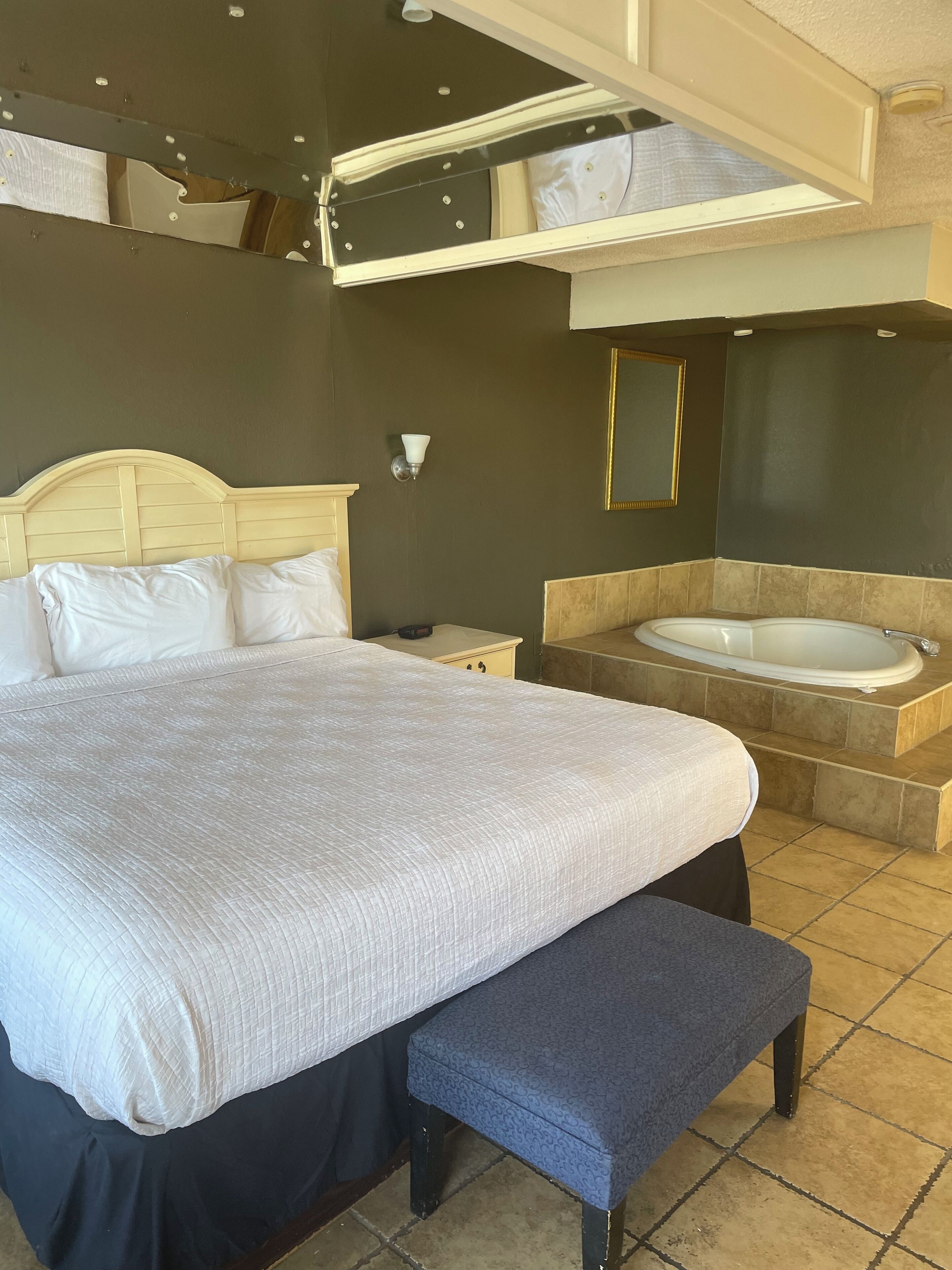 Een hotelkamer met een groot bed, een bankje, een nachtkastje, een wandlamp, een grote spiegel en een bad.