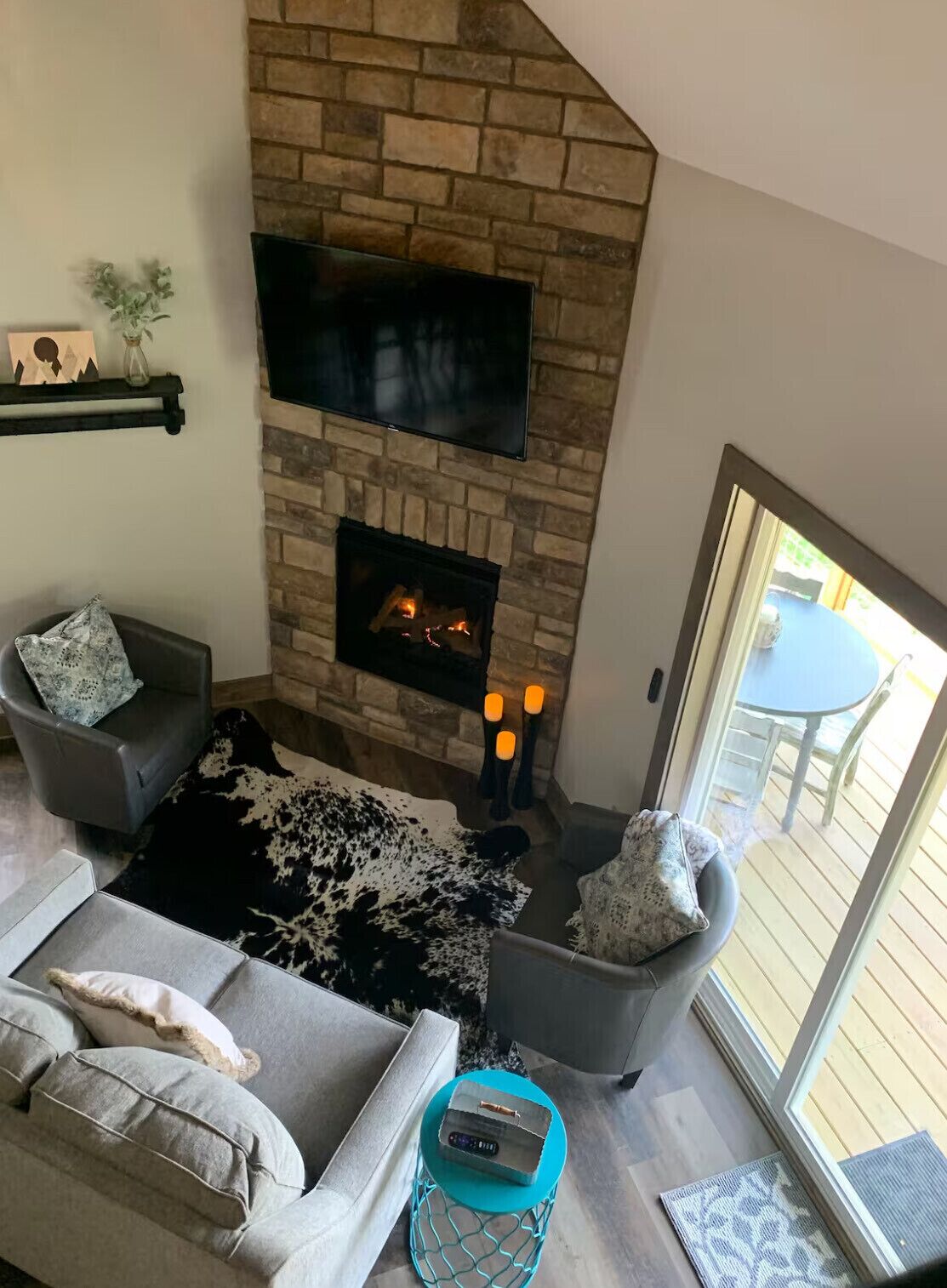 A cozy living room with a stone fireplace, a flat-screen TV mounted above it, a comfortable gray sofa with patterned cushions, and a black and white rug.