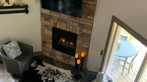 Un salon chaleureux avec une cheminée en pierre, un téléviseur à écran plat fixé au-dessus, un canapé gris confortable agrémenté de coussins à motifs et un tapis noir et blanc.