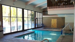 An indoor swimming pool with clear glass walls, a hot tub, and a wooden deck area.