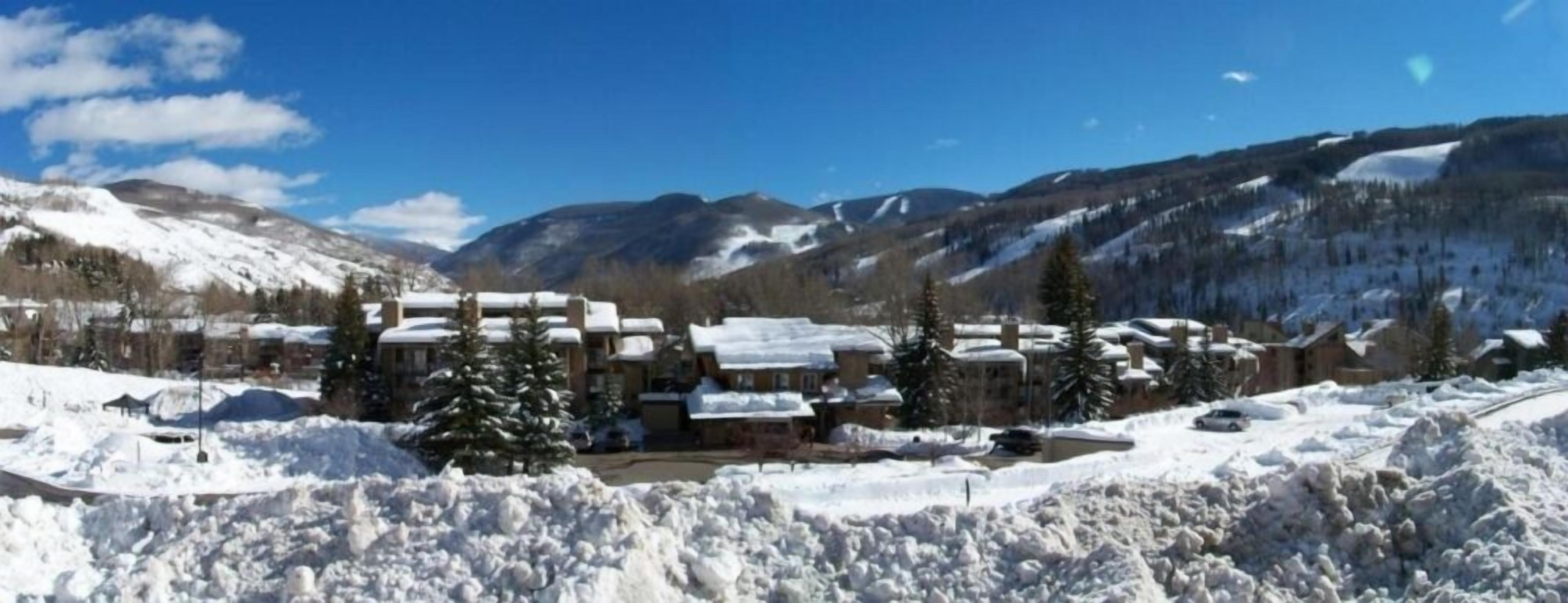 Une station de montagne enneigée, composée de plusieurs bâtiments, dont un chalet principal et des cabanes, le tout entouré de neige et d’arbres.