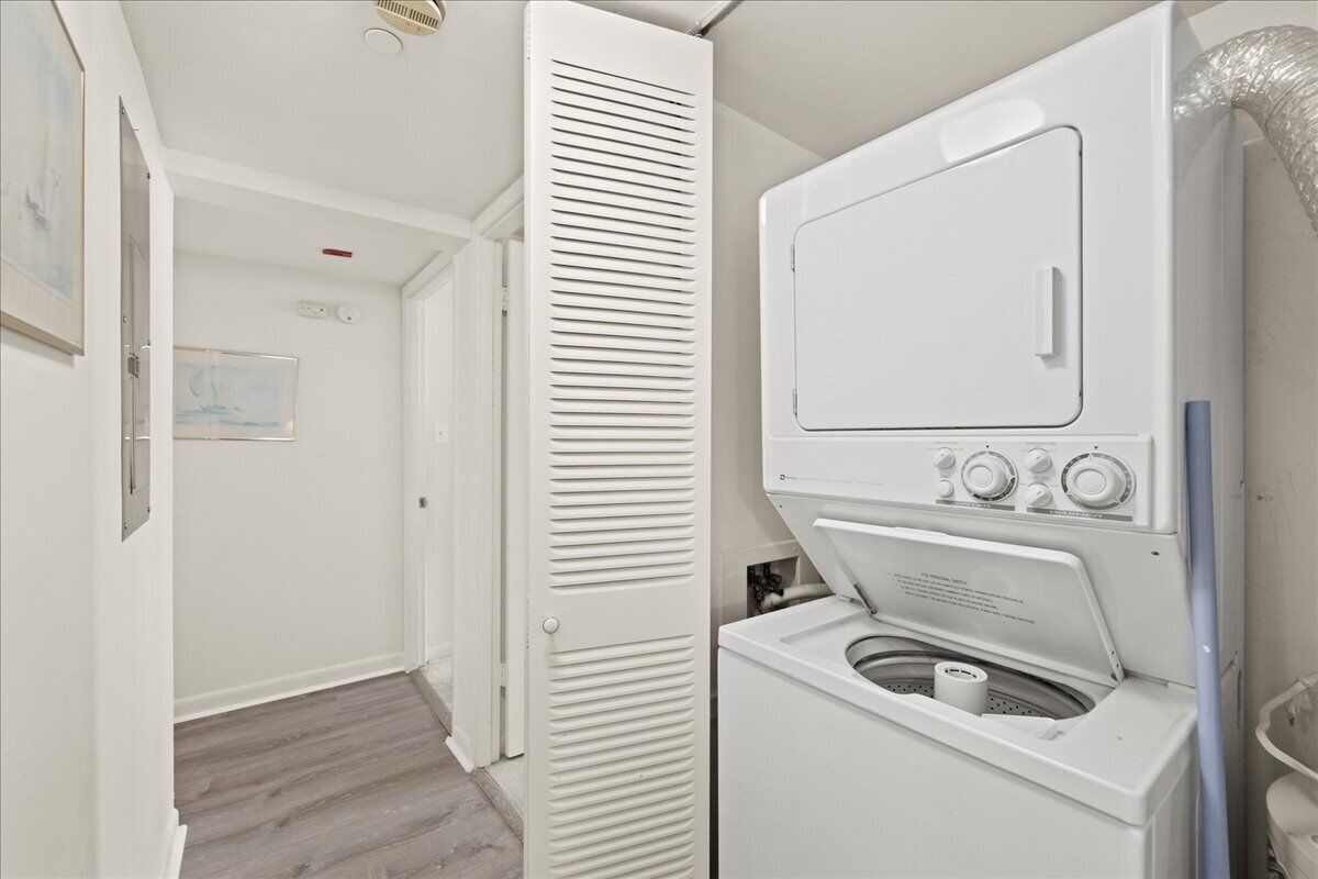 A laundry room with a stacked washer and dryer, a door with louvered panels, and a visible electrical outlet on the wall.