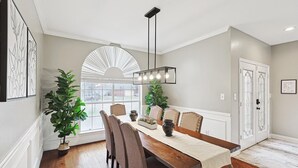 A dining room with a wooden table, six chairs, a chandelier, potted plants, and a large window with blinds. - Immaculate 5BR/2.5B vila with Pool and cinema (Irving)