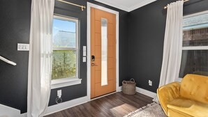 A room with a dark wall, a wooden door, a window with white curtains, a yellow sofa, and a white rug. - Luxury Comfort 5 Bedroom Home in Heart of Dallas (Dallas)